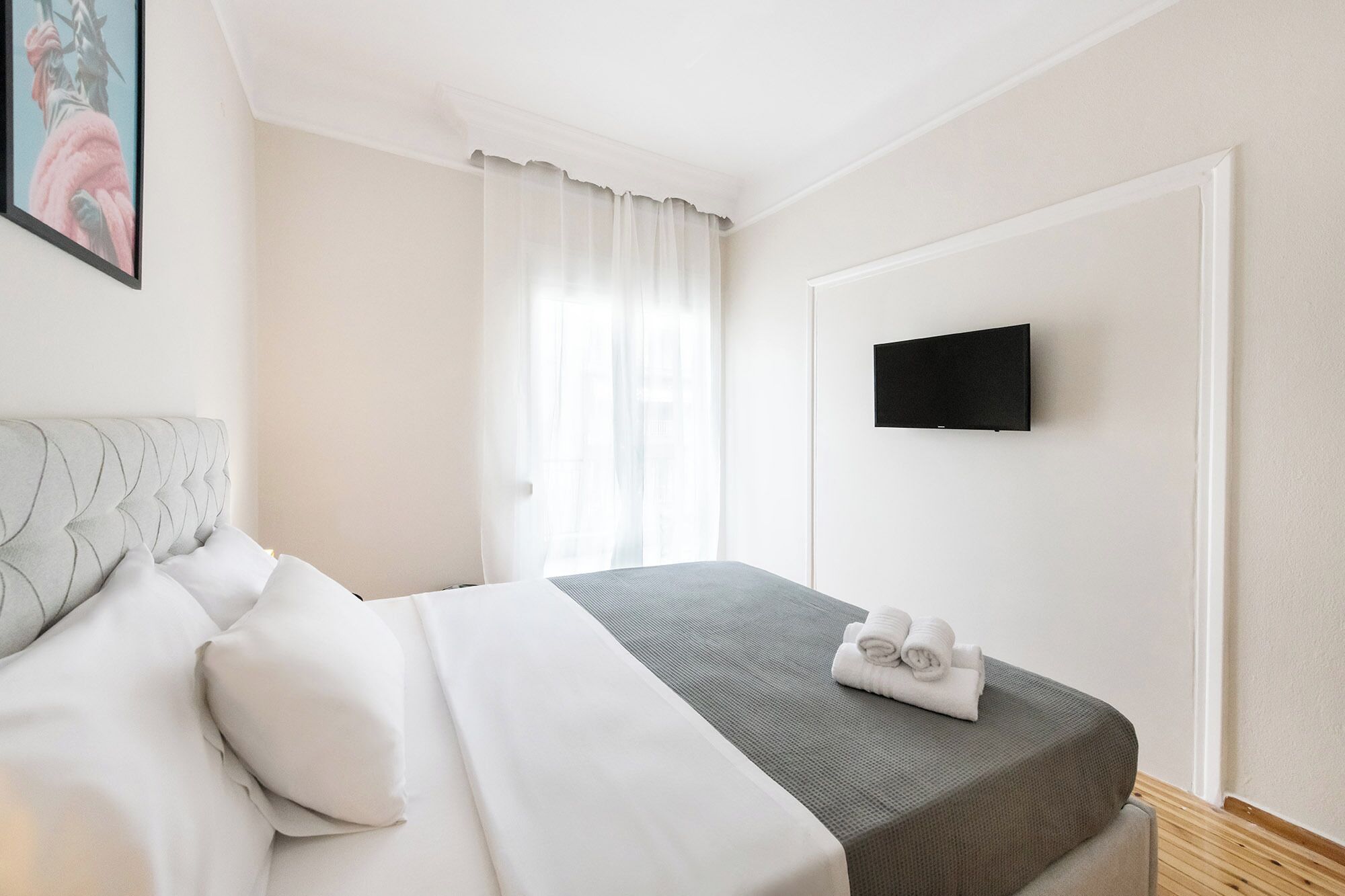 A hotel room with a bed, a flat-screen TV mounted on the wall, and a window with sheer curtains.
