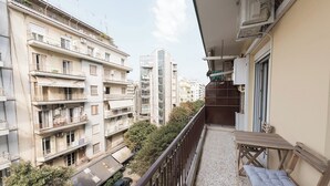 Ein Balkon mit Holztisch und gepolstertem Sitz, der auf eine Straße mit parkenden Autos und Gebäuden blickt.