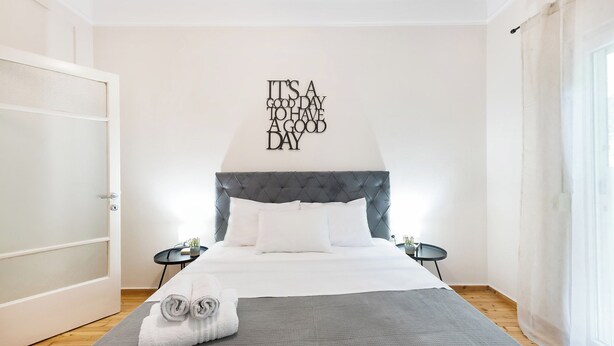 Ein ordentlich eingerichtetes Schlafzimmer mit einem gepolsterten Kopfende, einem Bett mit weißen Bettwäsche, einer grauen Tagesdecke und einem motivierenden Zitat an der Wand.