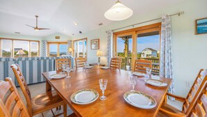Een eetruimte met een houten tafel gedekt voor zes personen, omringd door stoelen, en grote ramen met uitzicht op de oceaan.