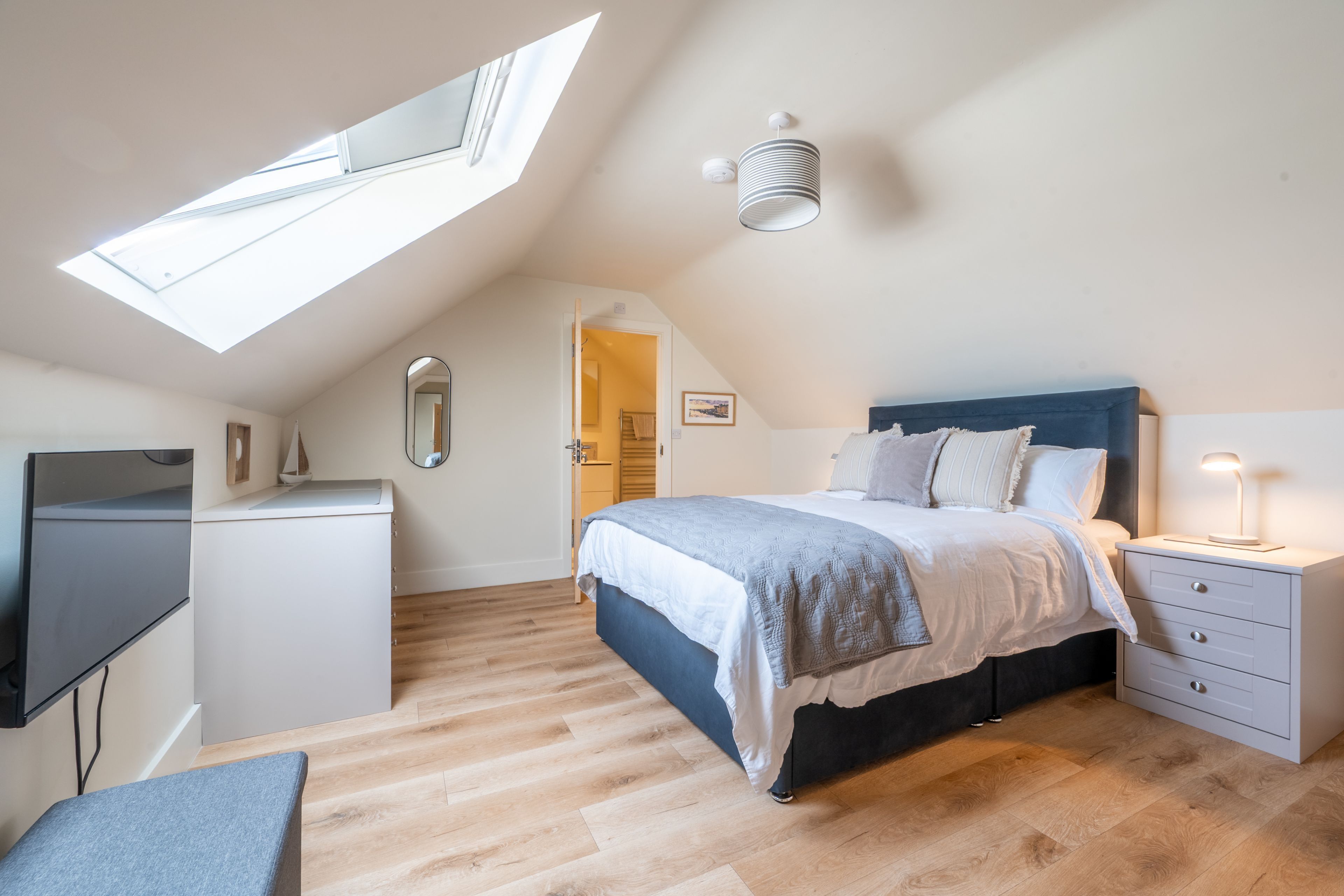 A bedroom with a skylight, a bed with a headboard, a bedside table with a lamp, and a TV on a stand.