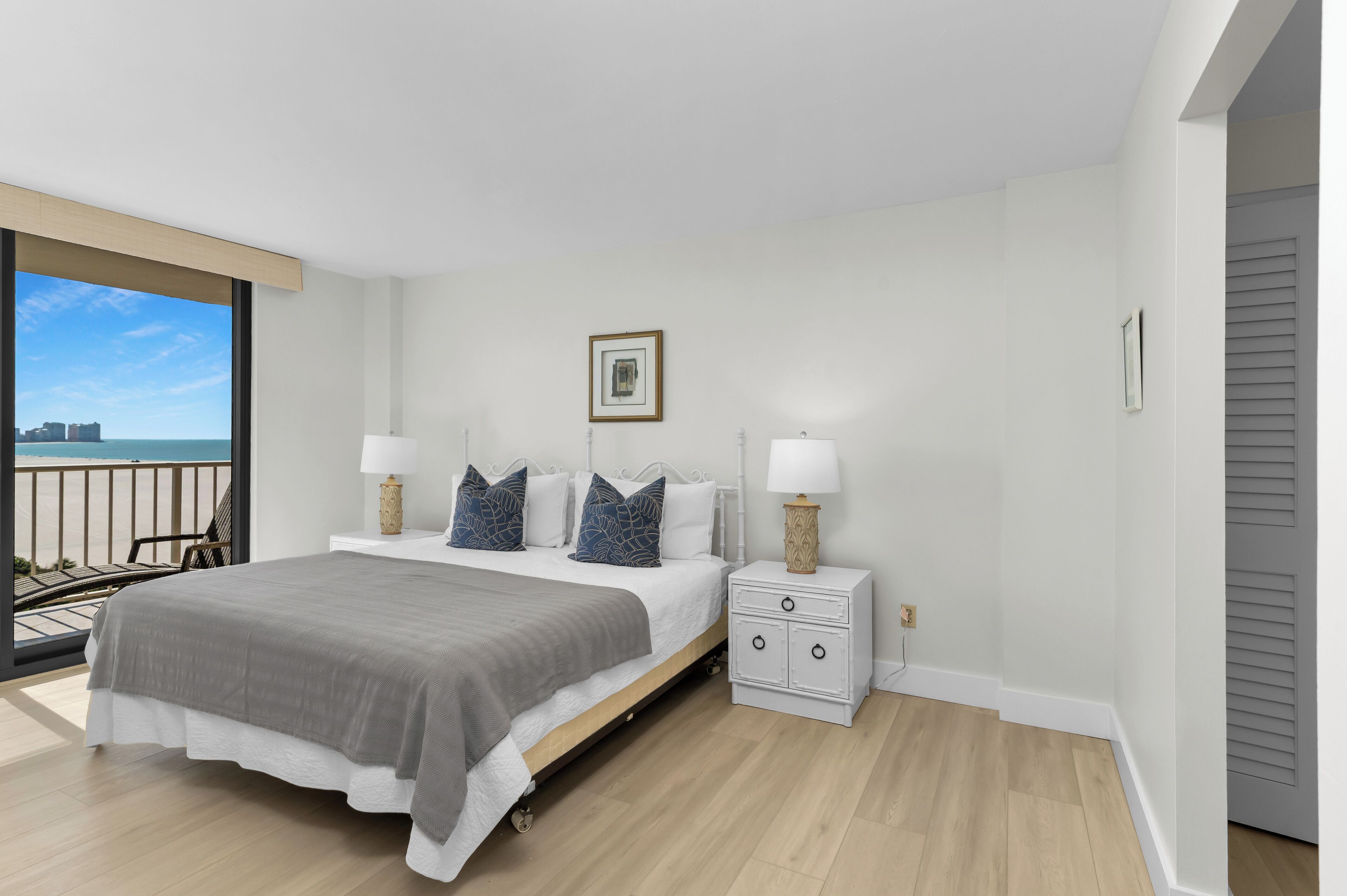 A modern bedroom with a large bed, bedside lamps, a small nightstand, and a view of the ocean.