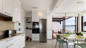 A modern kitchen with white cabinets, a stainless steel refrigerator, and a microwave. There is a dining area with a table and chairs, and a view of the outside through large windows. - Sea Breeze Jarea by Escape Home (Playa Blanca)