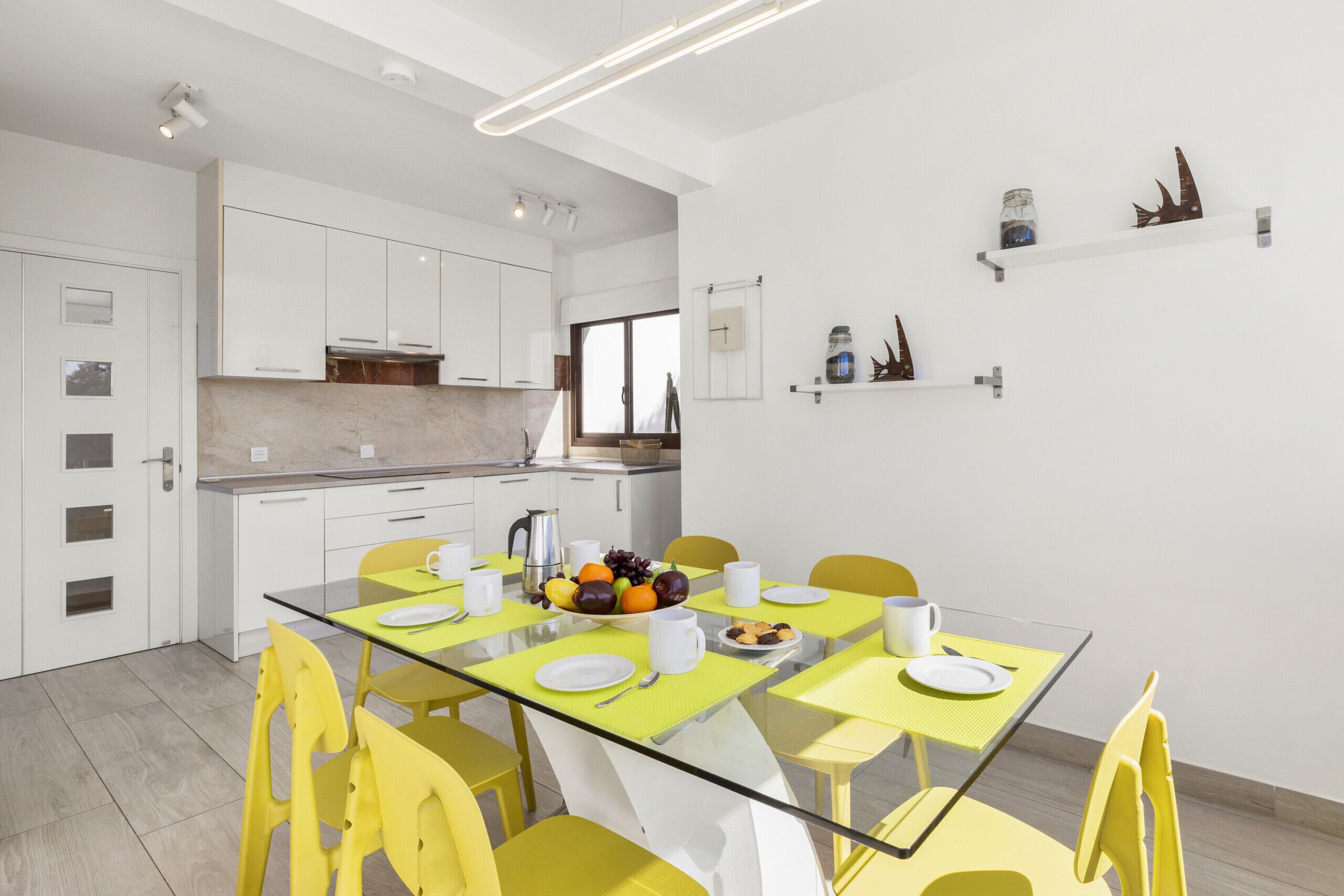 A modern kitchen with white cabinets, a glass dining table, yellow chairs, and a fruit bowl.