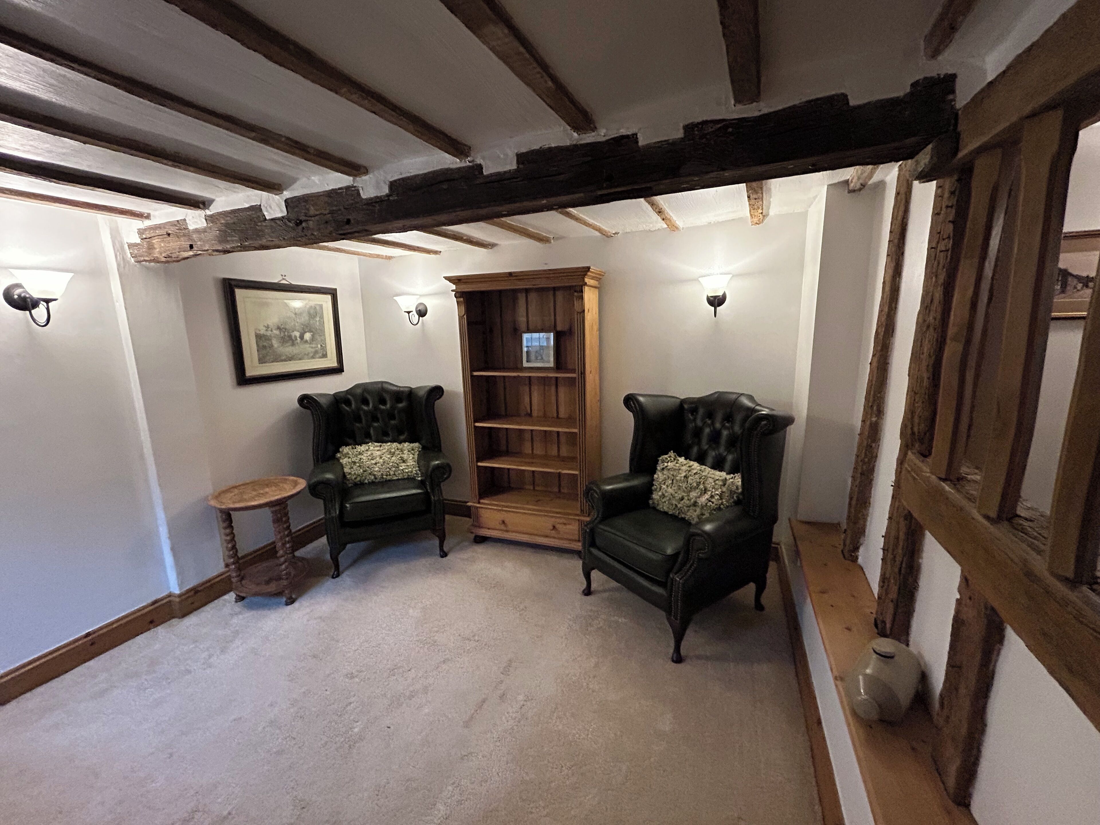 A room with exposed wooden beams, two leather armchairs with floral cushions, a small round table, and a bookshelf.