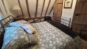 Un dormitorio con cama, vigas de madera, una mesita de noche, lámpara, puerta y un cuadro enmarcado en la pared.