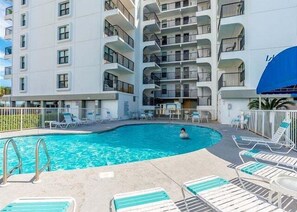 Ein mehrstöckiges Hotel mit Pool, Liegestühlen und einem blauen Sonnensegel.