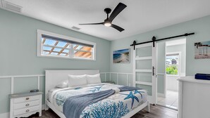 Une chambre à coucher comprenant un lit, une table de chevet, une commode, un ventilateur de plafond, une fenêtre avec des stores et une porte donnant sur une autre pièce.