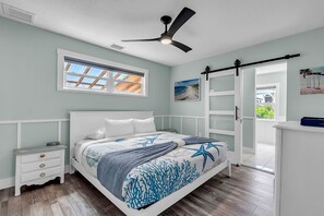 A bedroom with a bed, a nightstand, a dresser, a ceiling fan, a window with blinds, and a door leading to another room.