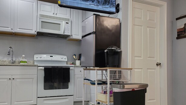 Una cocina moderna con armarios blancos, un refrigerador de acero inoxidable, un microondas y un horno. Hay un televisor montado sobre la estufa, y se ve un bote de basura.