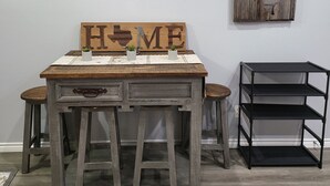 Una habitación con una mesa de madera, dos taburetes, un aire acondicionado instalado en la pared, un letrero decorativo de madera y un estante metálico negro.
