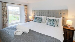 Una cama bien hecha con edredón gris y blanco, dos almohadas y una toalla blanca enrollada. Hay dos mesitas de noche con lámparas, una ventana con cortinas florales y vistas a zonas verdes.
