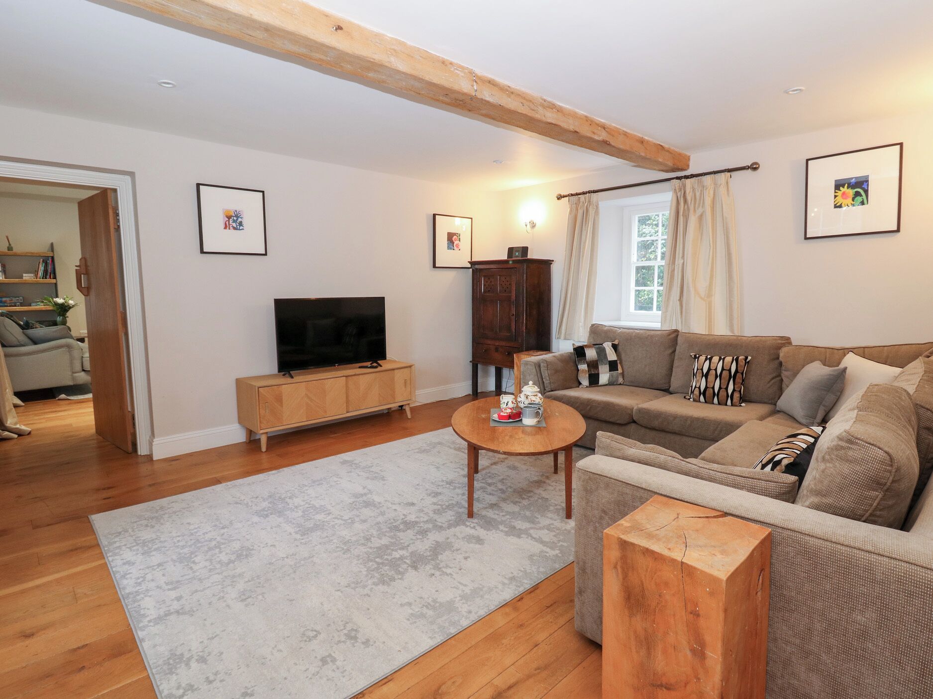Una sala de estar con piso de madera, un sofá color beige, un televisor sobre un mueble de madera y una ventana con cortinas.