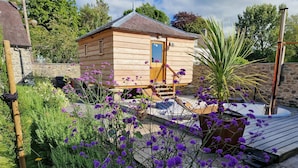 Una cabaña de madera con un pequeño porche, rodeada de un jardín con flores moradas y una planta de palmera.