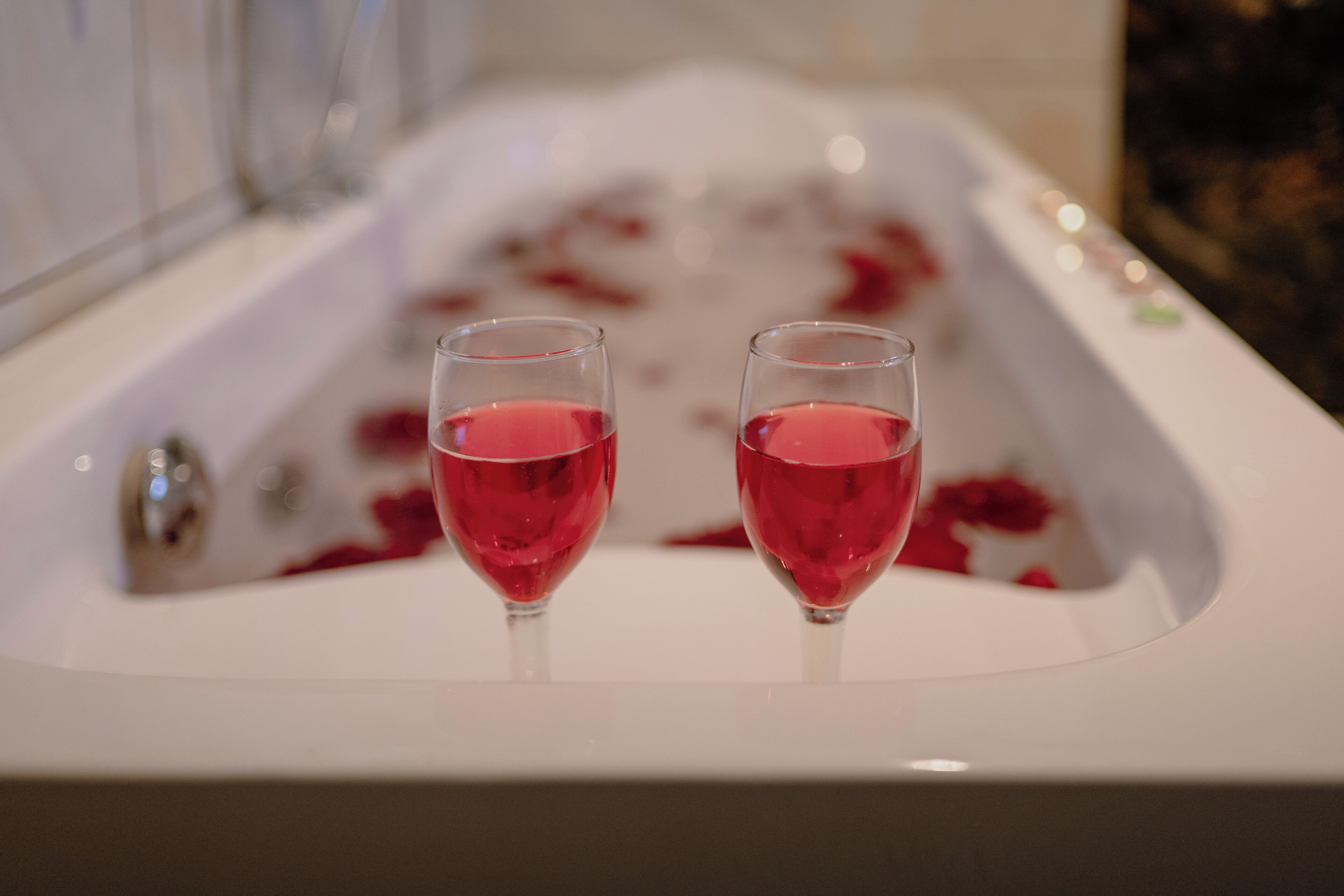 Una bañera llena de agua y pétalos de rosa, con dos vasos de líquido rojo en el borde.