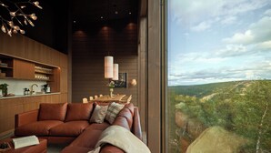 A modern living room with a large brown leather sofa, a wooden dining table with chairs, and a view of a green landscape through large windows.