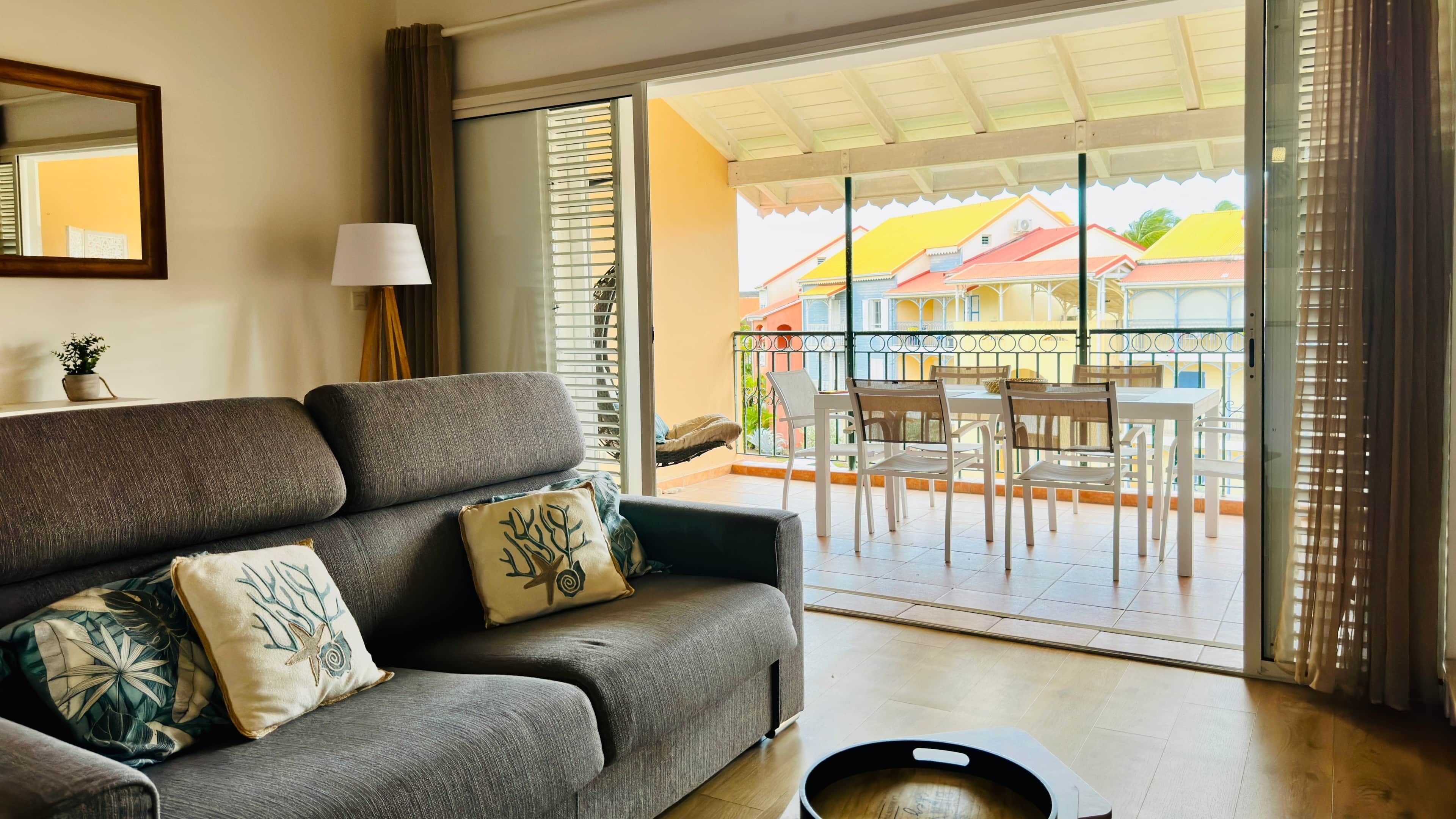 A living room with a grey sofa, a small table with a lamp, and a view of a balcony with outdoor furniture.