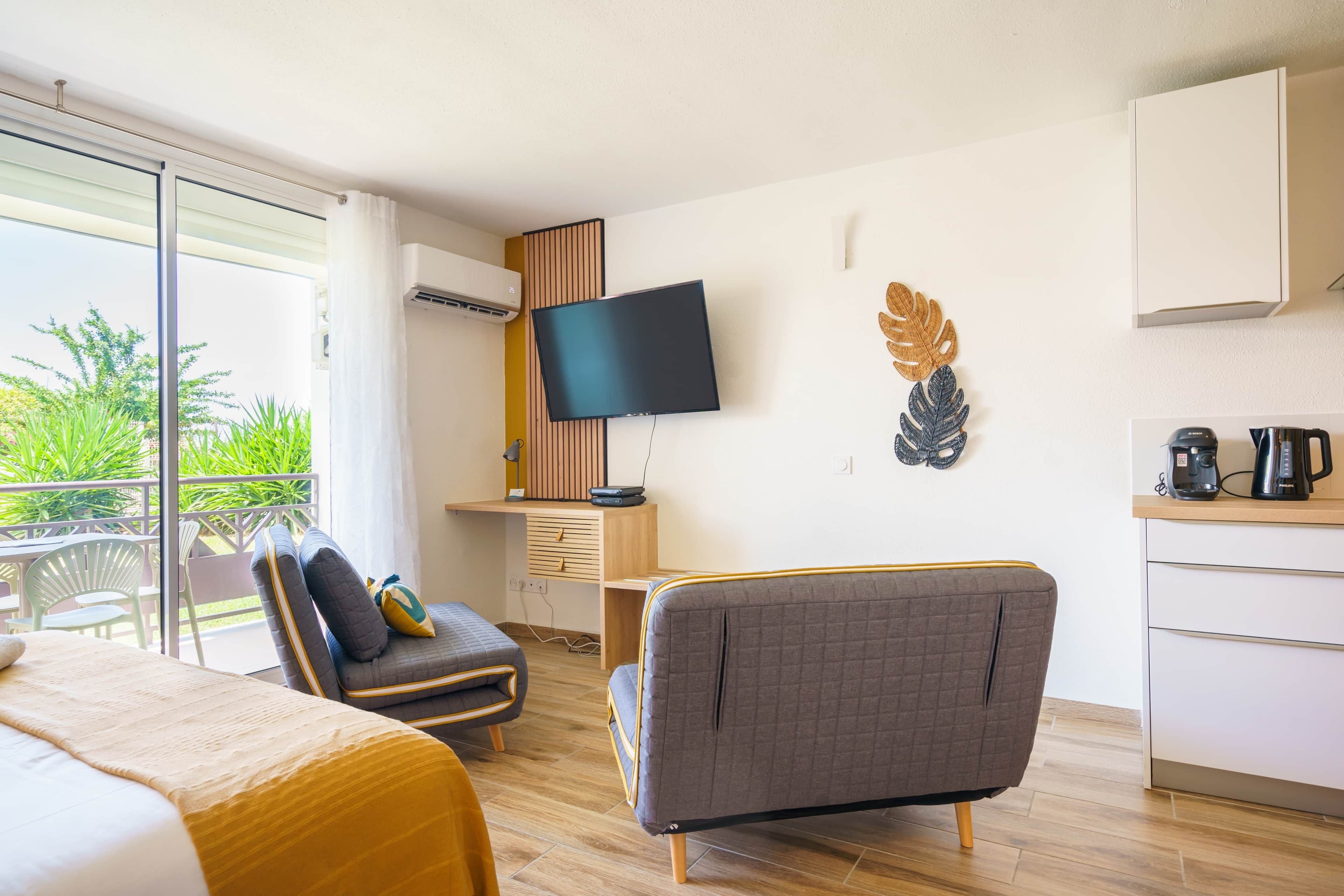 A modern hotel room with a bed, a sofa, a TV, a desk, and a balcony with plants.
