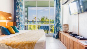 Quarto de hotel com uma cama, uma mesa de cabeceira de madeira, uma televisão na parede e uma varanda com mesa e cadeiras.