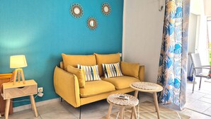 A cozy living room with a mustard sofa, two side tables, a lamp, and decorative wall art. - Comfortable studio with terrace, cistern and pool access (Le Gosier)