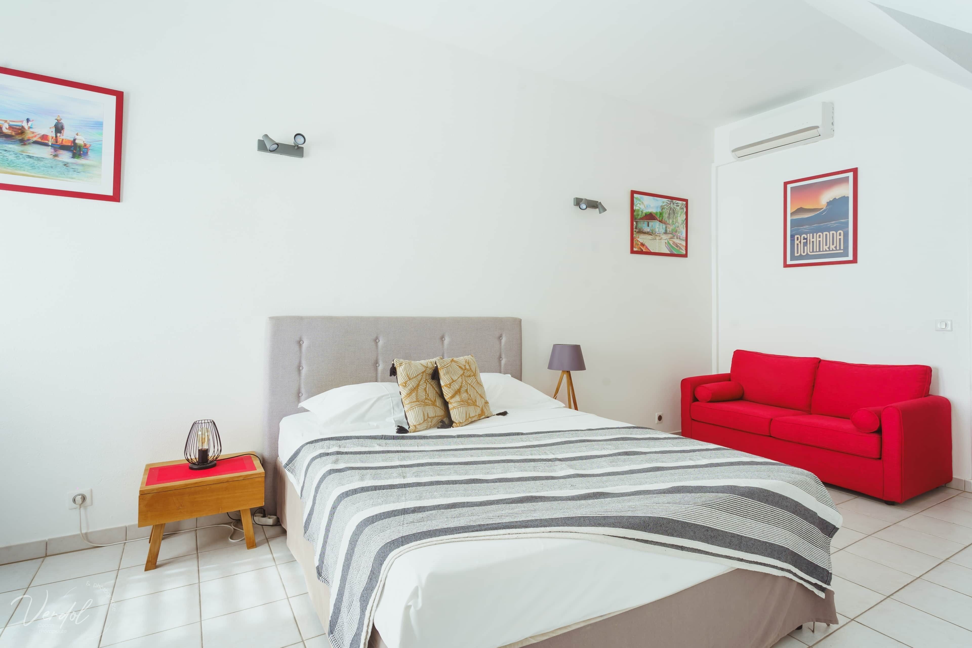 A bedroom with a bed, a red sofa, a nightstand, and two framed pictures on the wall.
