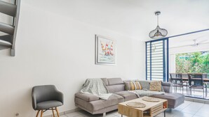 A modern living room with a grey sofa, a wooden coffee table, and a grey armchair. There is a painting of sailboats on the wall and a dining area with a table and chairs visible through a glass door. - Spacious duplex near the beach with cistern (Le Gosier)