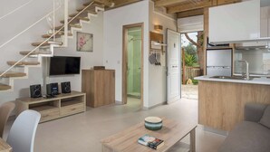 Een moderne woonkamer met een televisie, speakers en een salontafel. Er is een trap die naar een andere kamer leidt, en een keukenhoek met een spoelbak en kasten.