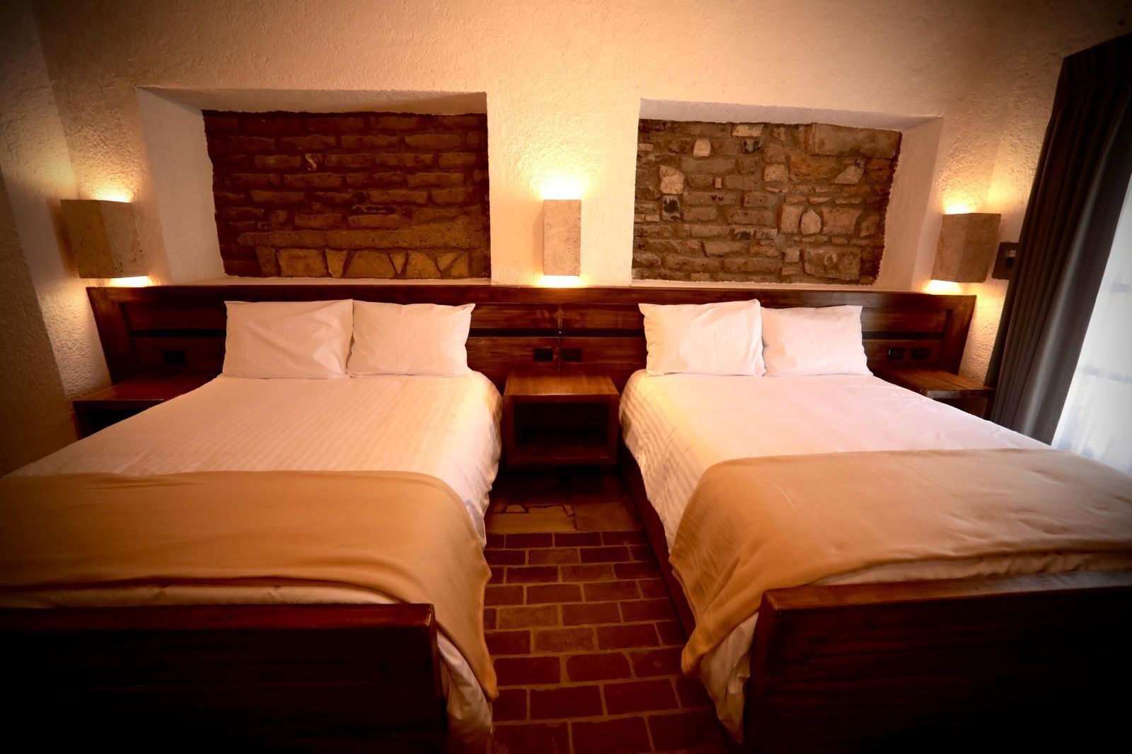 A hotel room with two beds, a brick floor, stone wall decor, and wall-mounted lamps.