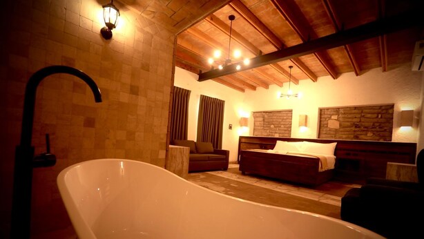 A room with a large bathtub, wooden ceiling beams, a bed with a headboard, and a stone wall. - Hotel Villa de la Parra (Parras de la Fuente)