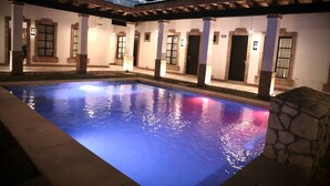 A swimming pool with a stone wall and a covered patio area with columns and doors. - Hotel Villa de la Parra (Parras de la Fuente)