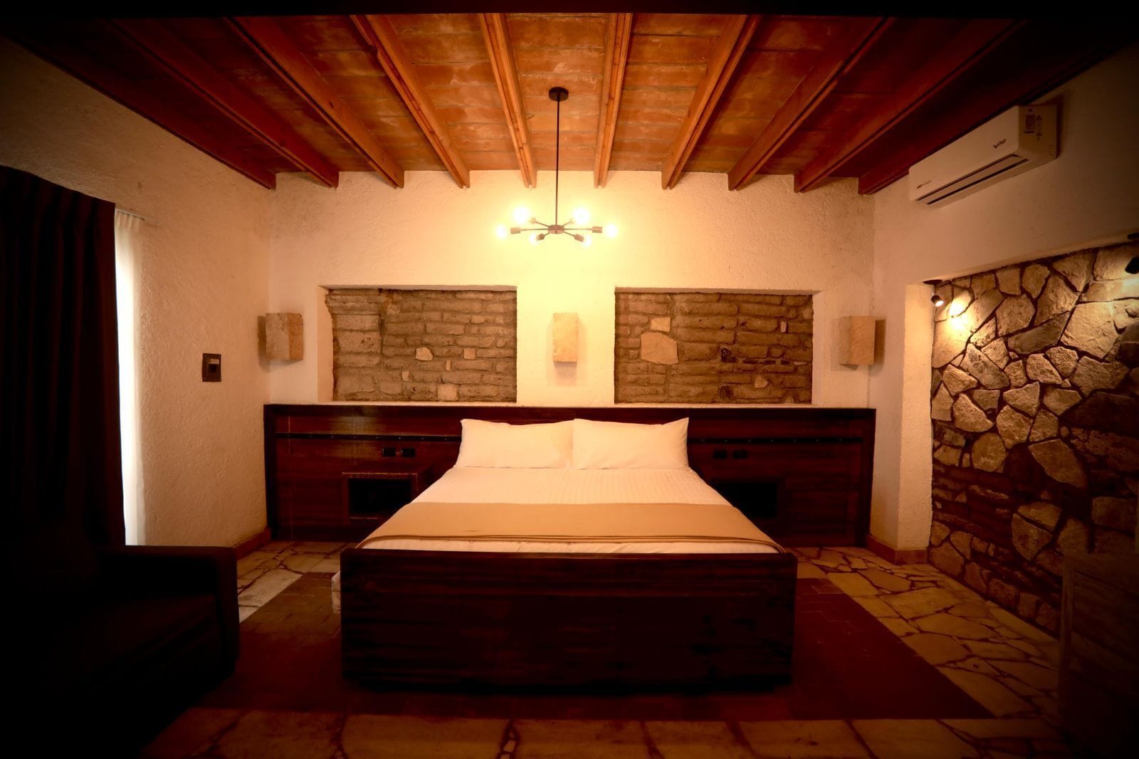 A bedroom with a stone wall, wooden ceiling beams, a bed, and a stone fireplace.
