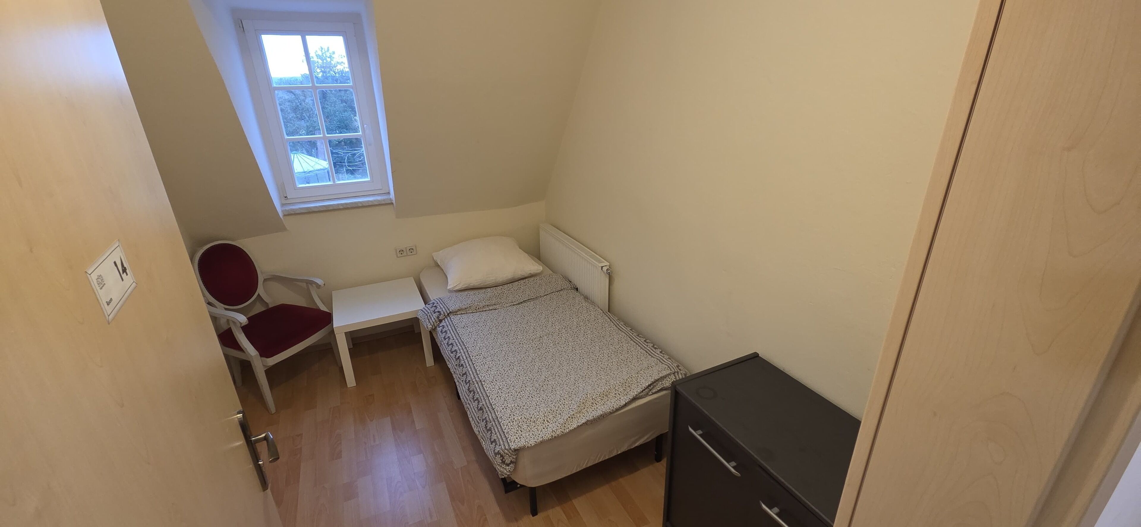 A small, single-room bedroom with a bed, a chair, a small table, a window, and a wardrobe.