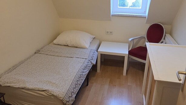 A small, single-room apartment with a sloped ceiling, a bed, a desk, a chair, and a window providing natural light.
