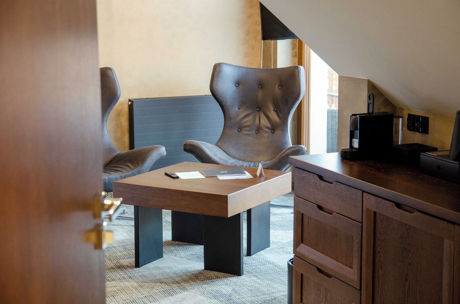 A modern room with a dark brown desk, two leather chairs, a coffee machine, and a large window.