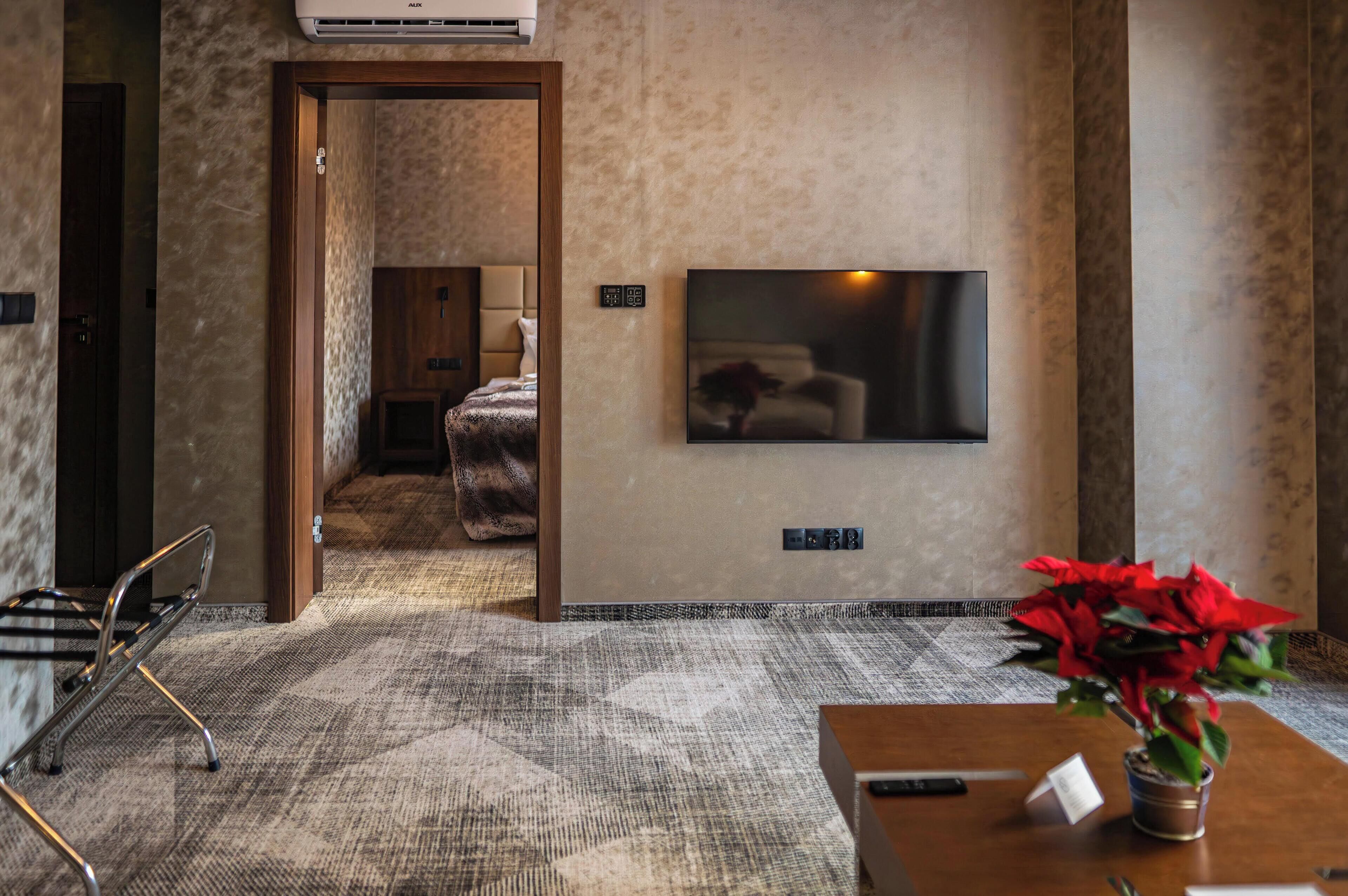 A hotel room with a large flat-screen TV mounted on the wall, a wooden door leading to another room, a patterned carpet, and a small table with a red flower vase.