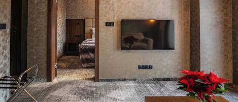 A hotel room with a large flat-screen TV mounted on the wall, a wooden door leading to another room, a patterned carpet, and a small table with a red flower vase.