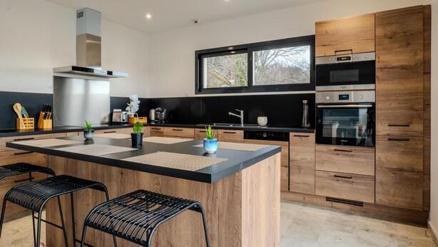 A modern kitchen with a central island, black countertops, wooden cabinets, and stainless steel appliances.