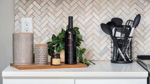 A kitchen with a herringbone tile backsplash, white cabinets, and a countertop with a plant, a bottle, and utensils. - Backstage Bungalow- Cozy 3BR Retreat in The Nations + Near Downtown (Nashville)