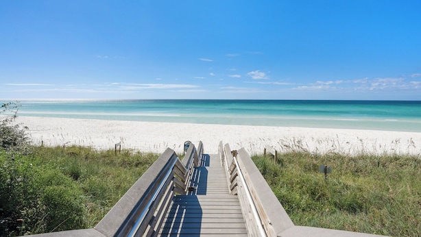 Een houten looppad dat naar een strand leidt met helderblauw water en wit zand.