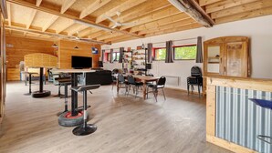 A spacious room with a high ceiling, wooden beams, a dining area with chairs and a table, a bar area with a counter, and a television mounted on the wall.