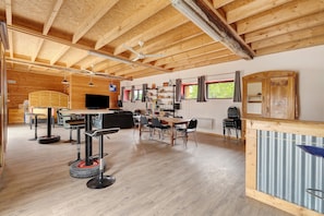 A spacious room with a high ceiling, wooden beams, a dining area with chairs and a table, a bar area with a counter, and a television mounted on the wall.