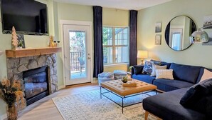 A living room with a stone fireplace, a flat-screen TV mounted above it, a blue sofa, a coffee table, and a large mirror on the wall. - Chalet 113 at the Official Collins Lake Resort (Government Camp)