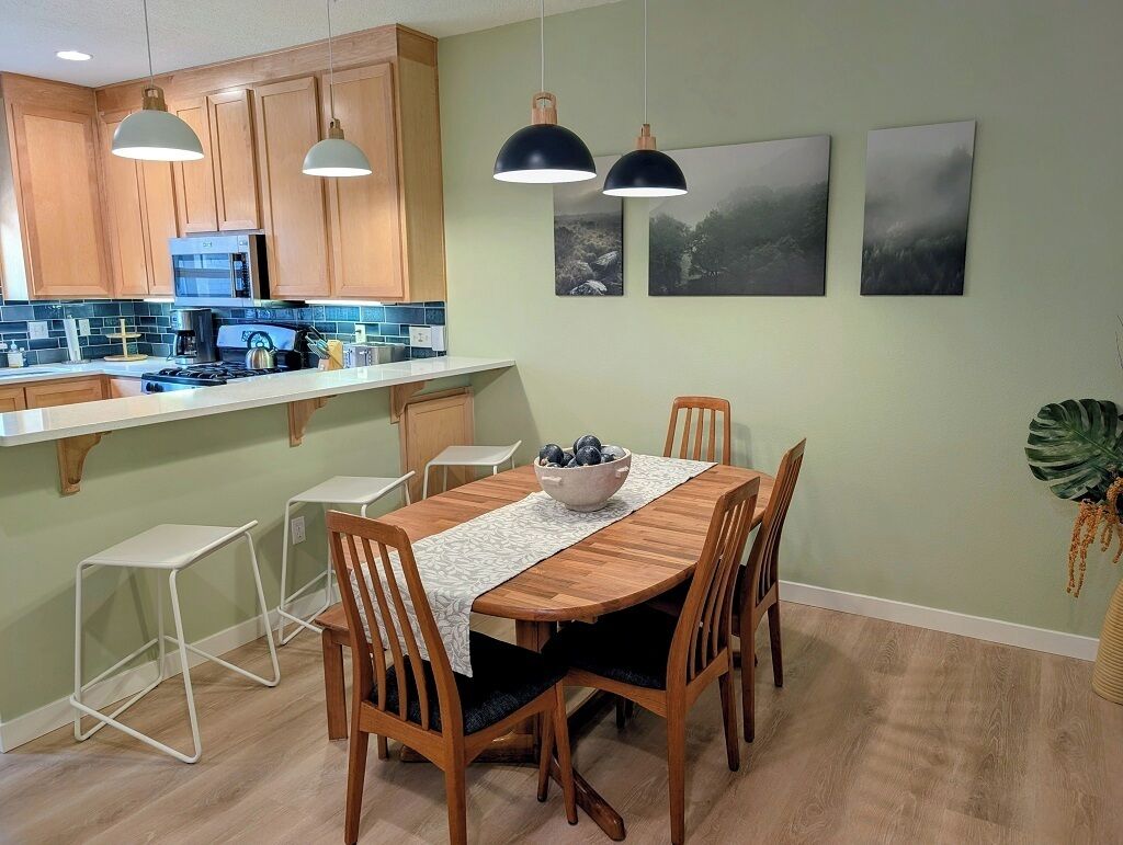 A modern kitchen with wooden cabinets, a central island, and pendant lighting. There are two framed pictures on the wall and a potted plant to the side.