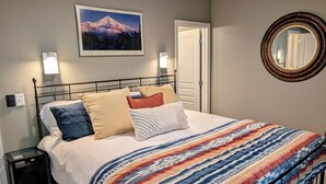 A neatly made bed with a colorful striped blanket, a framed picture of a mountain on the wall, and a round mirror on the wall. - Chalet 113 at the Official Collins Lake Resort (Government Camp)