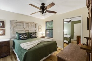 Ein Schlafzimmer mit Bett, Deckenventilator, Fenster mit Jalousien, Kommode und Spiegel.