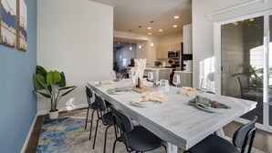Een moderne eetruimte met een lange tafel gedekt voor vier personen, omringd door zwarte stoelen. Daarboven hangt een kroonluchter en links is een potplant te zien.