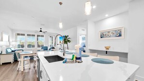 A modern kitchen with a large island, white cabinetry, and a dining area with blue chairs. - The Grove Juniper 228 - 2 Bedrooms - Lazy River - Seascape Resort (Miramar Beach)