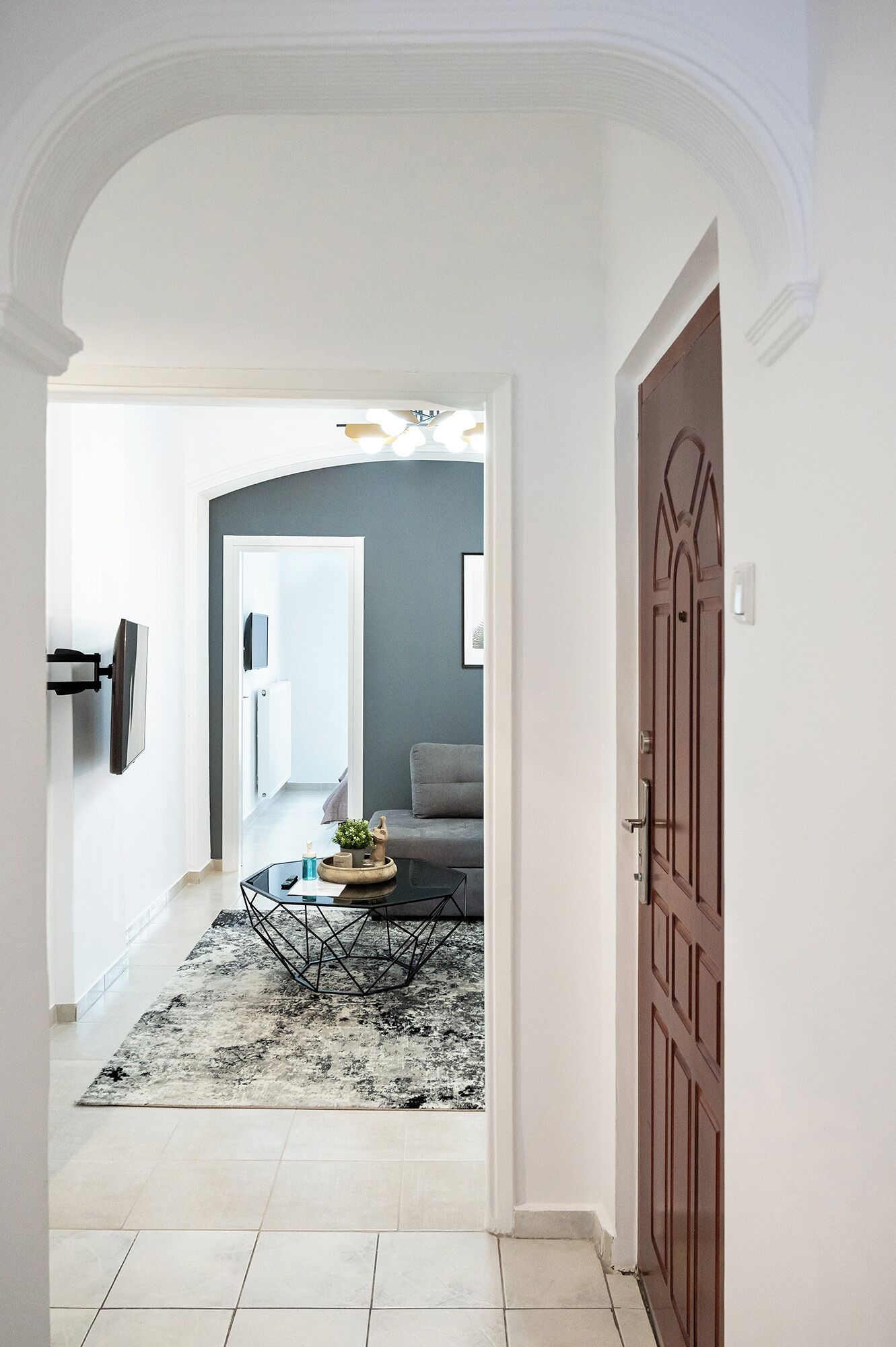 A hallway with a tiled floor, a black and white patterned rug, a glass coffee table with decorative items, a grey sofa, and a wooden door.