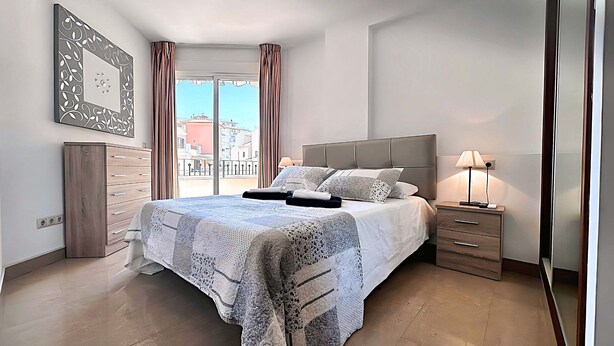A bedroom with a bed, bedside table, lamp, and a view of buildings through the window. - Euromar 121 (Torrox)