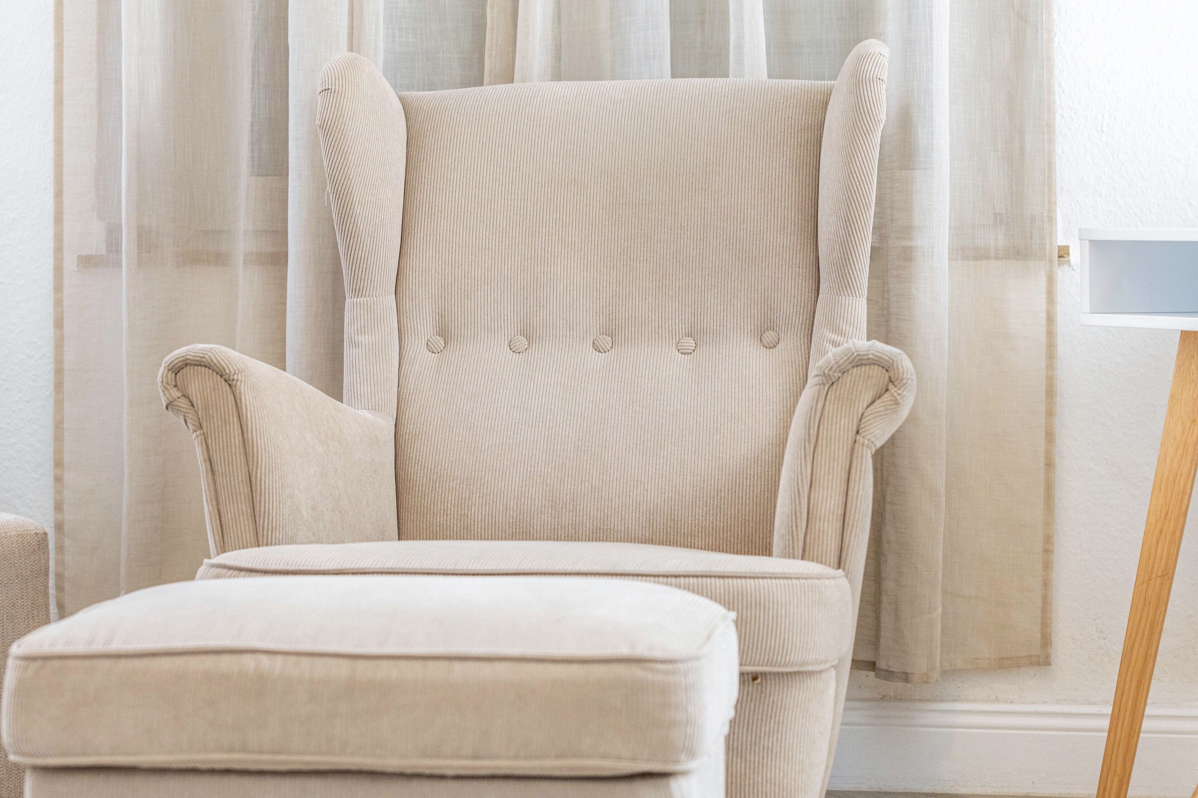 Ein beiges Polsterarmchair mit hoher Lehne und passendem Hocker in einem Raum mit transparenten Vorhängen und einer weißen Wand.